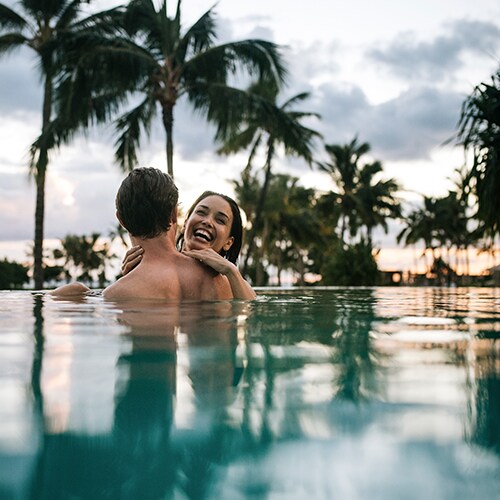 Two smiling adults in an infinity pool, with palms silhouetted along the shoreline