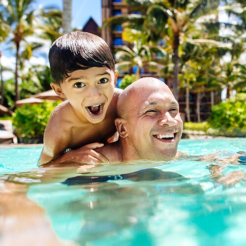 In sparkling blue water, a mother and father, his young son perched on his back, smile as they enjoy swimming together
