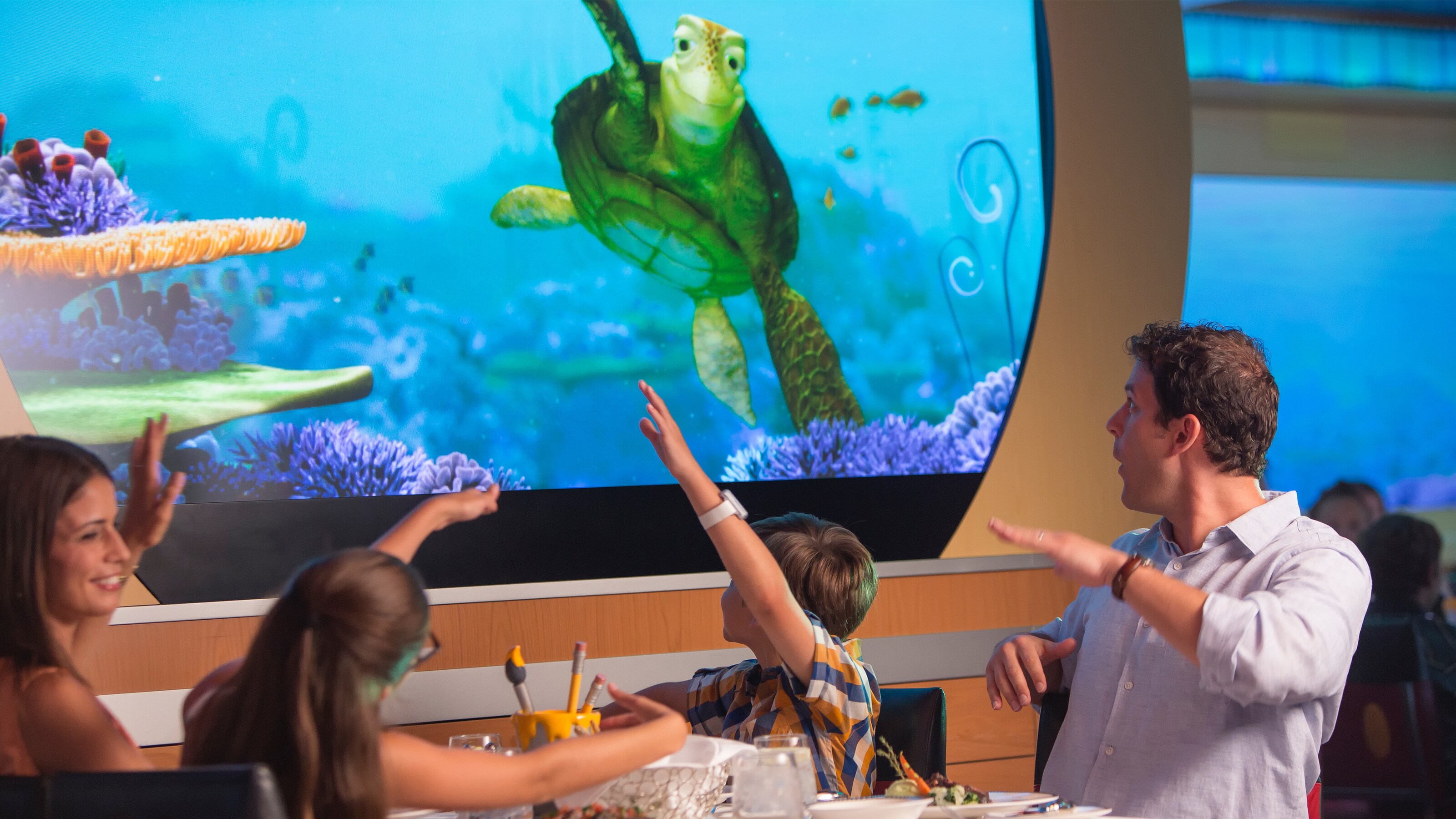 Una familia de cuatro integrantes mira una tortuga marina animada en una pantalla digital mientras cena en Animator's Palate en un crucero de Disney