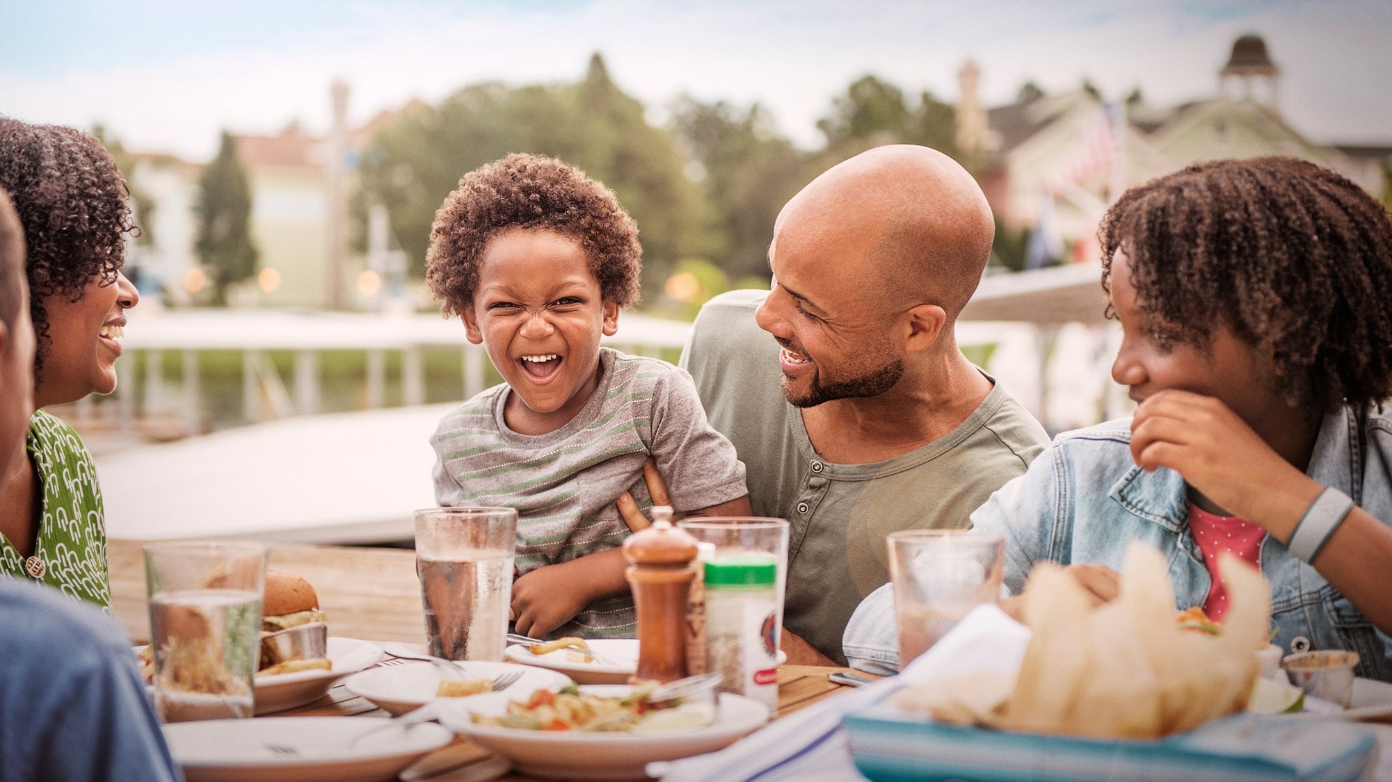 A bald man holds a young boy making a silly face while eating with his family at an outdoor table 