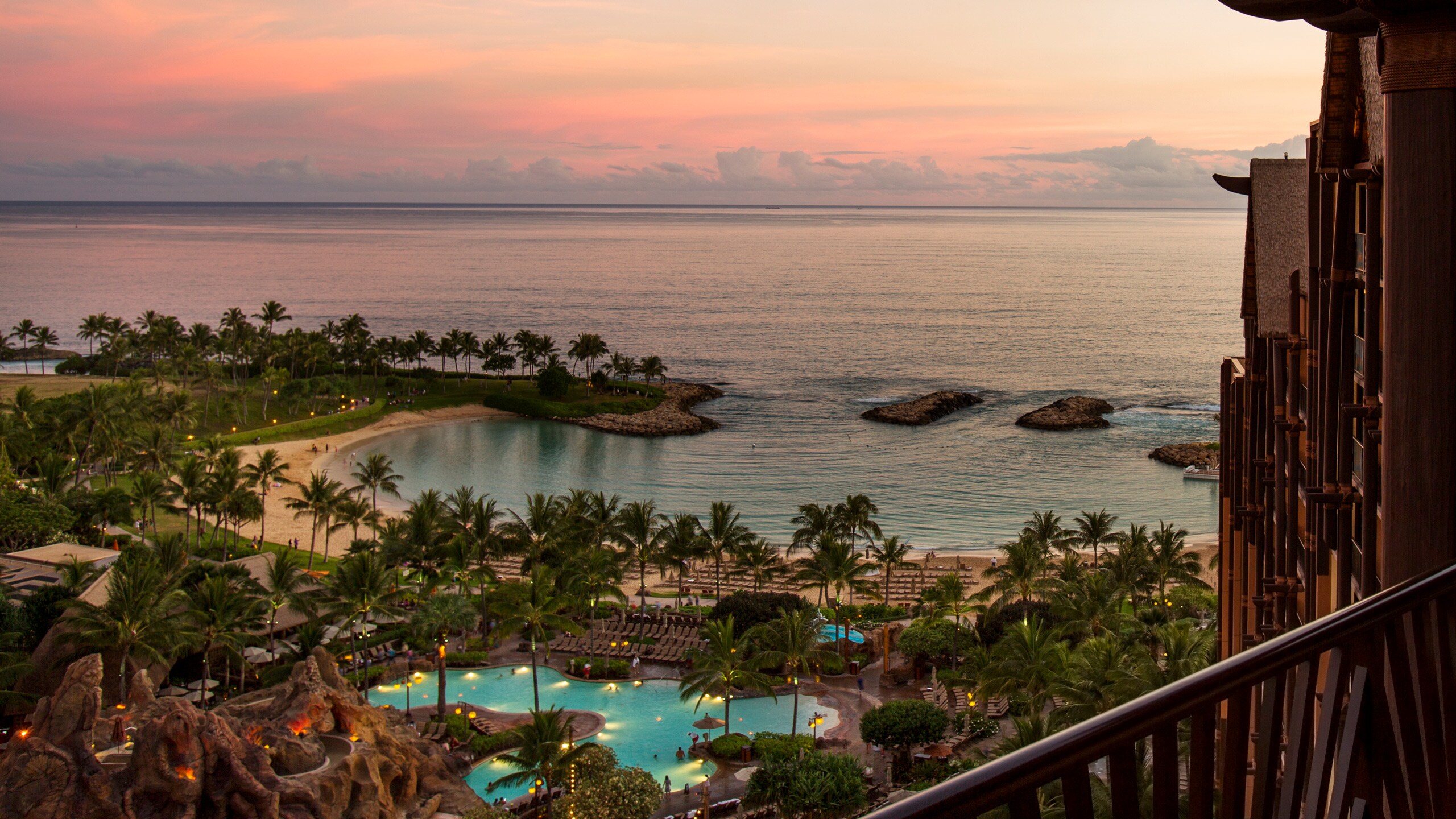 A sunset view from a high balcony of a pool surrounded by palm trees and a calm lagoon