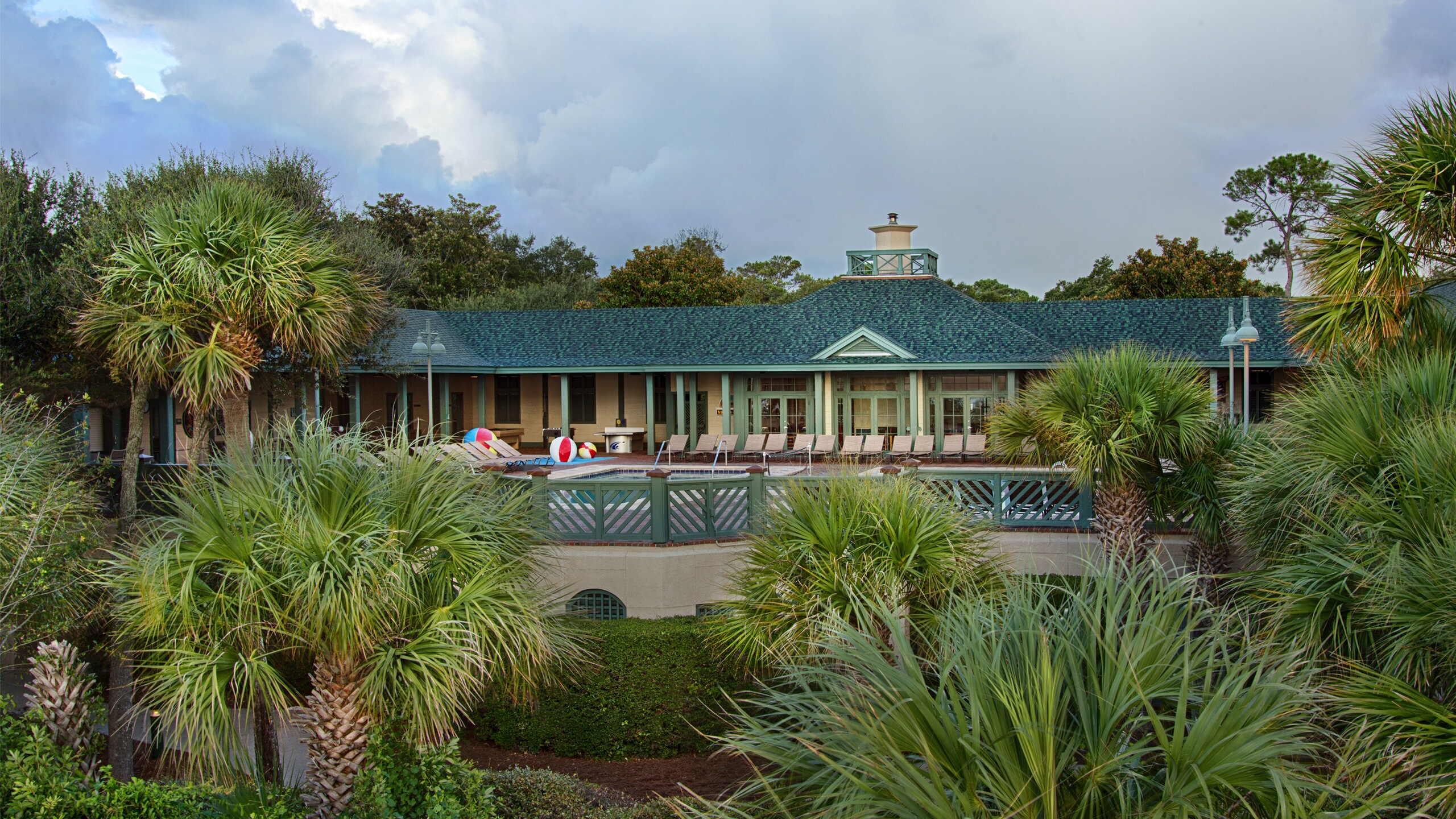 A pool and patio area with a 1 story beach house surrounded by a fence, bushes and palm trees