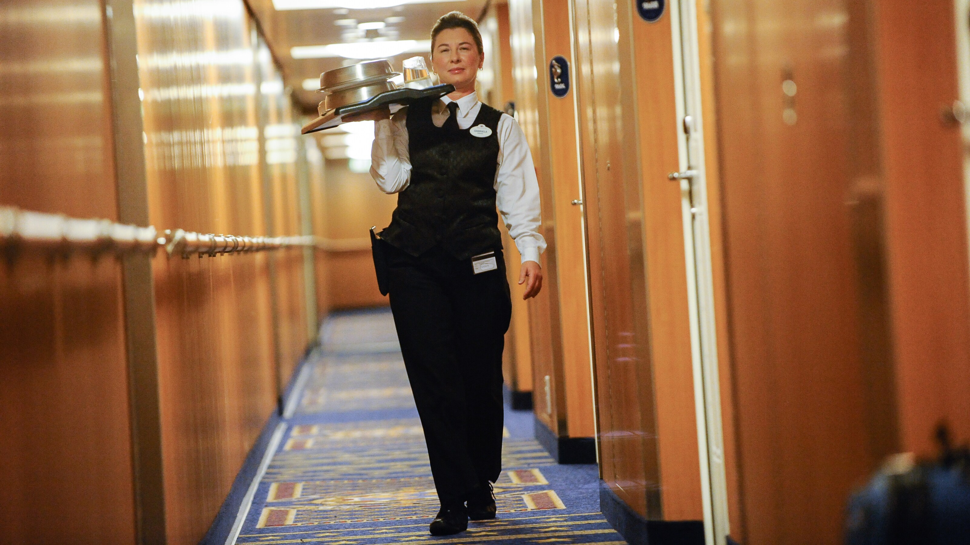 A Cast Member walks down a hallway holding a room service tray with metal plate covers and drinks