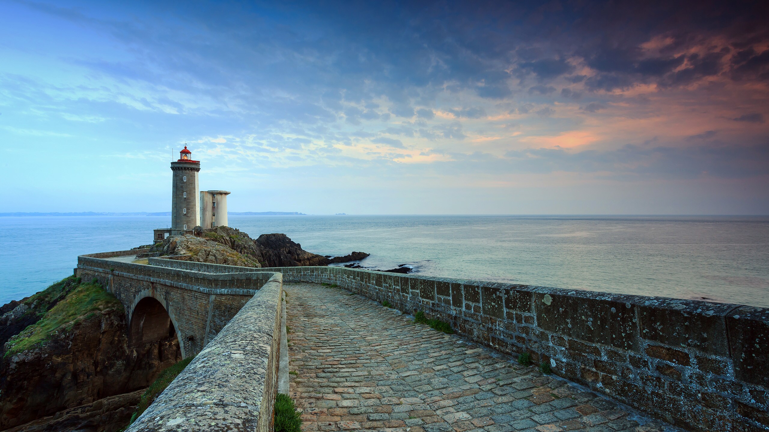 A stone walkway and bridge near a lighthouse and the ocean