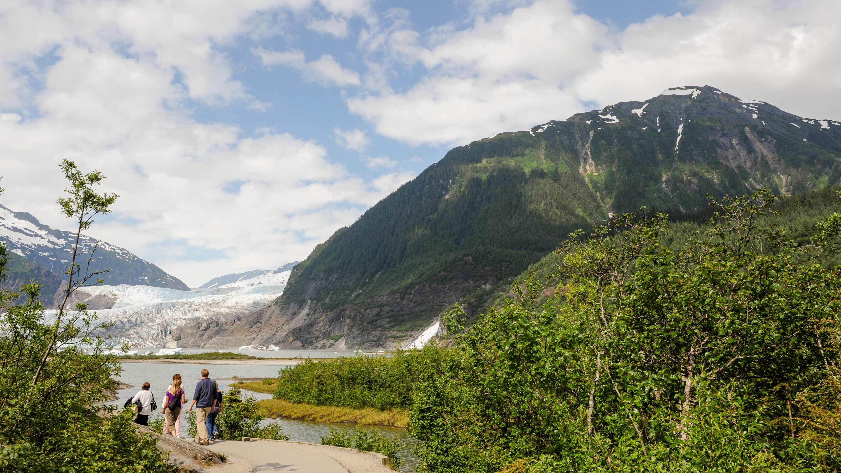 A small group of people look toward a glacier across a lake and bordered by mountains