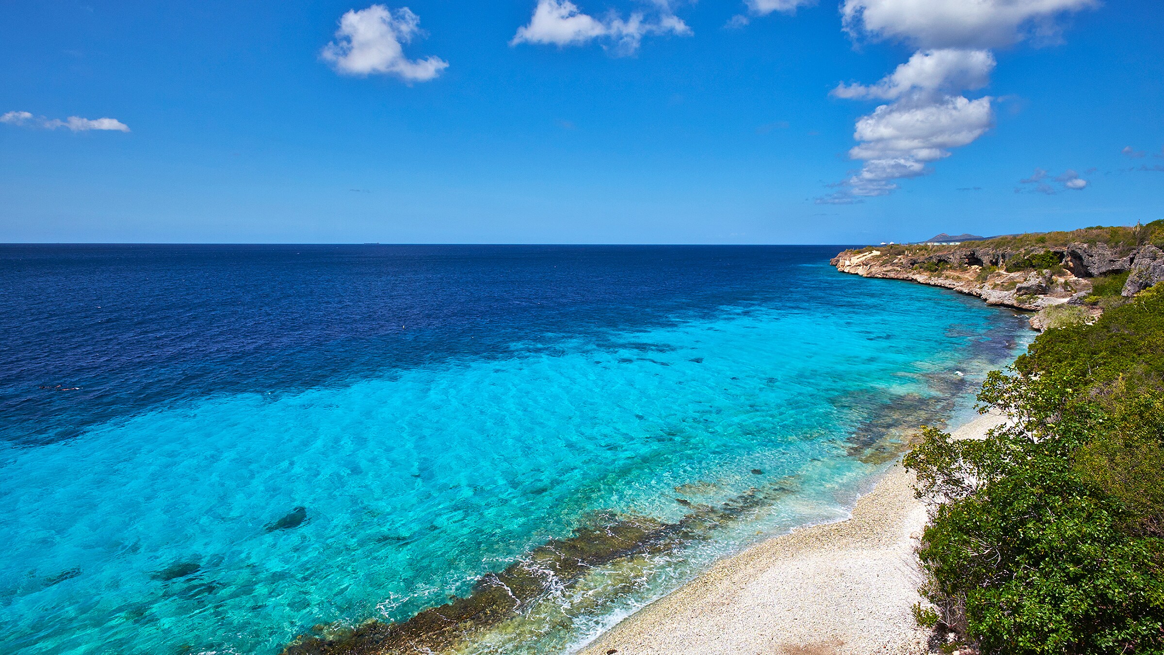 A pristine shallow water beach with a thin strip of sand against a lush and craggy shoreline