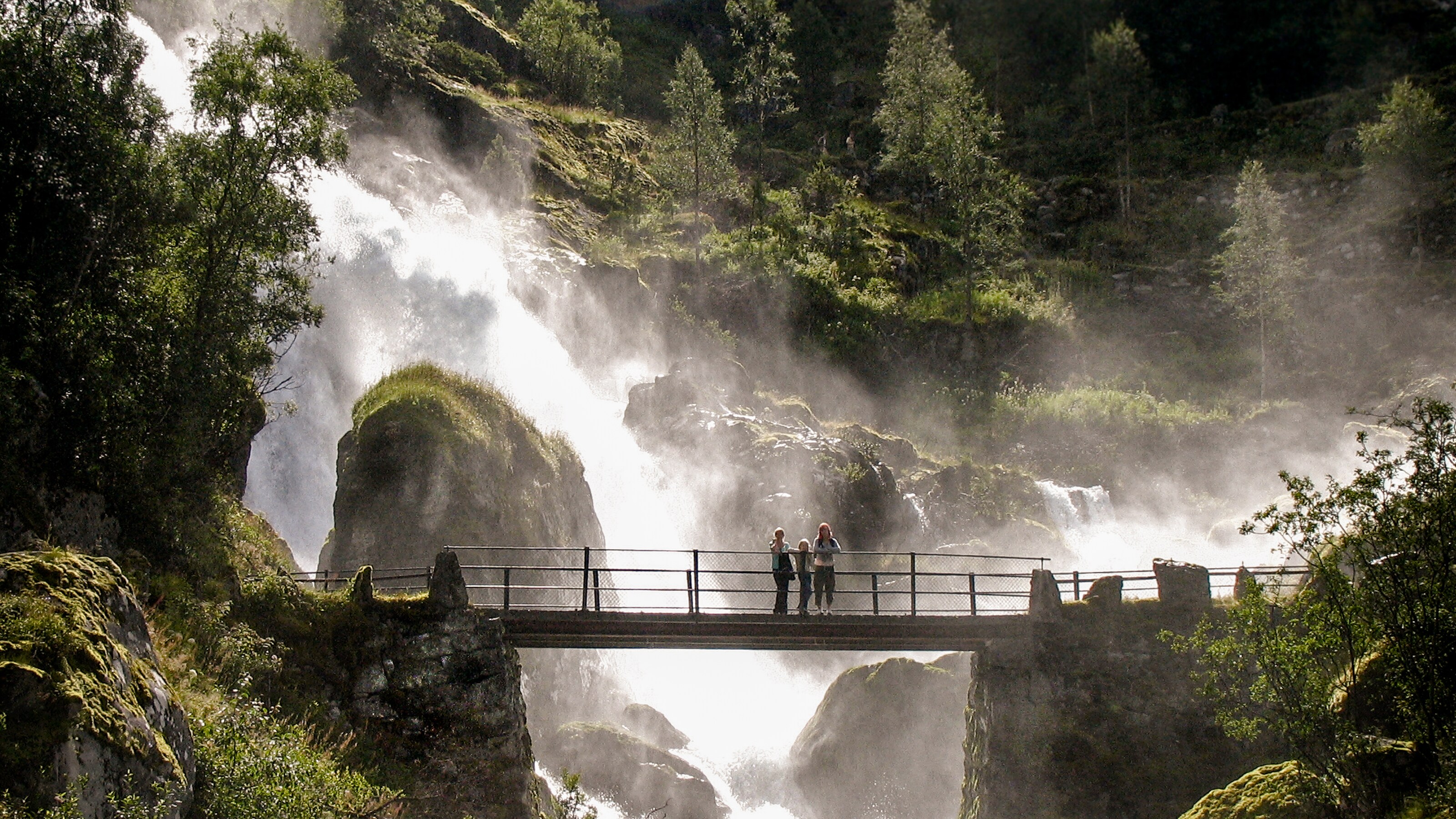 Three people stand on a bridge looking out with a waterfall cascades down rocks behind them
