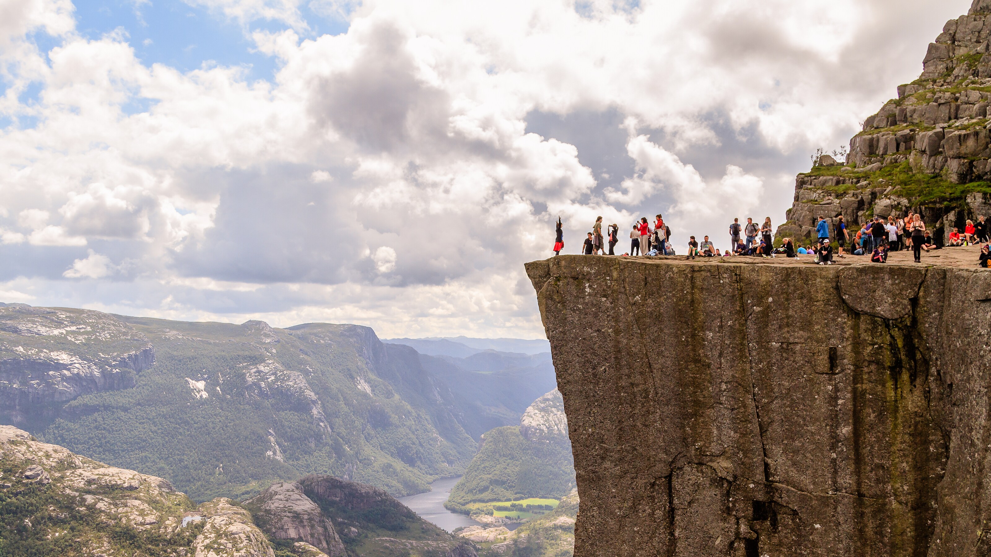 An assortment of people sitting and standing above a sheer cliff overlooking a valley