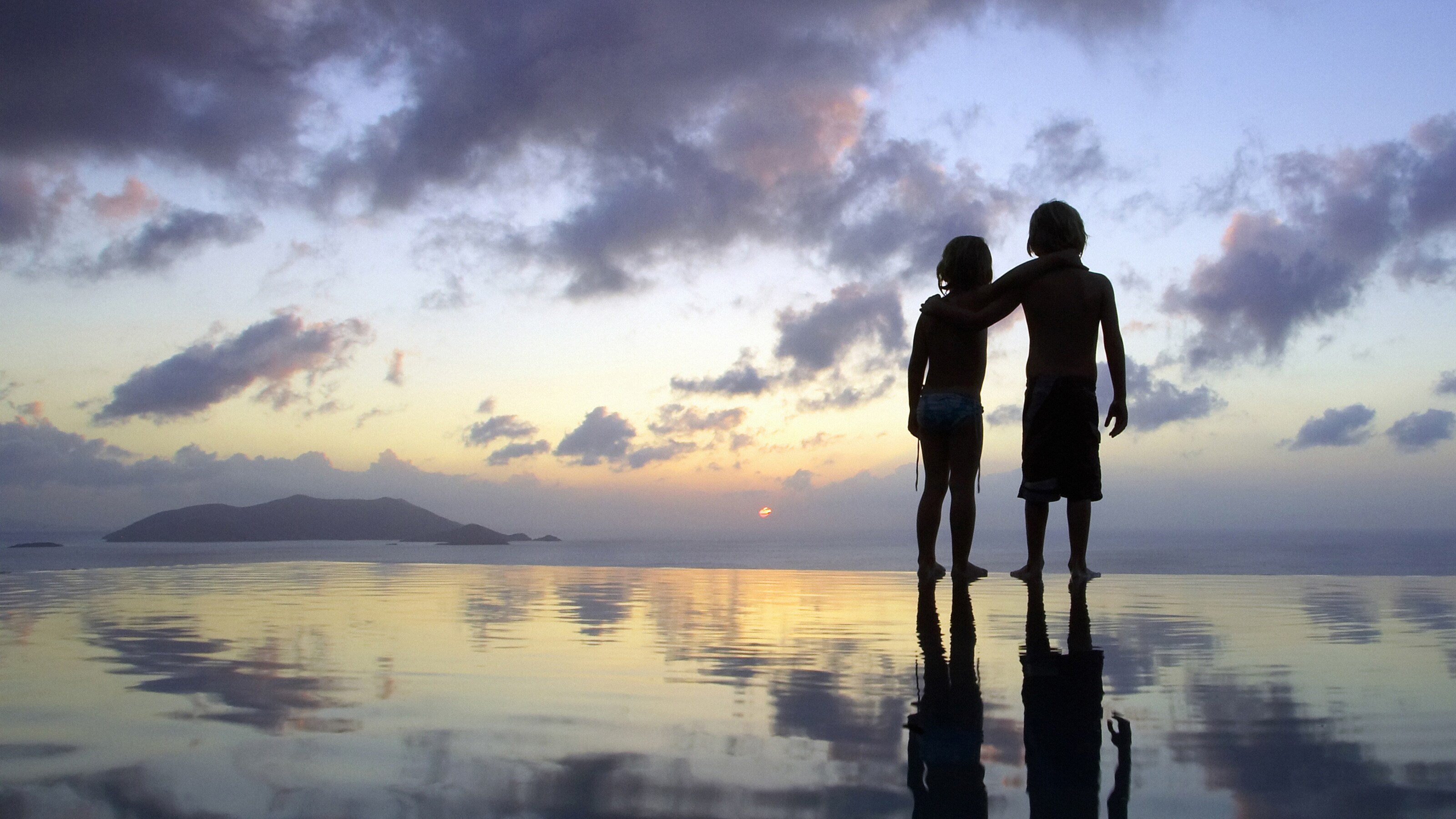A boy puts his arm around a girl, as they walk in silhouette on the shoreline at sunset