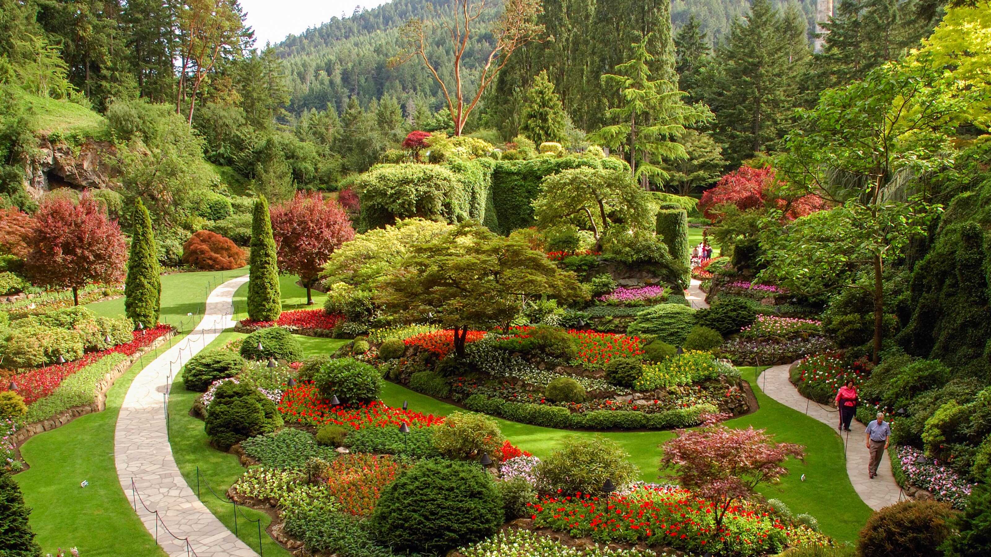 A stone path encircles a garden populated by arranged flowers grass paths and trees