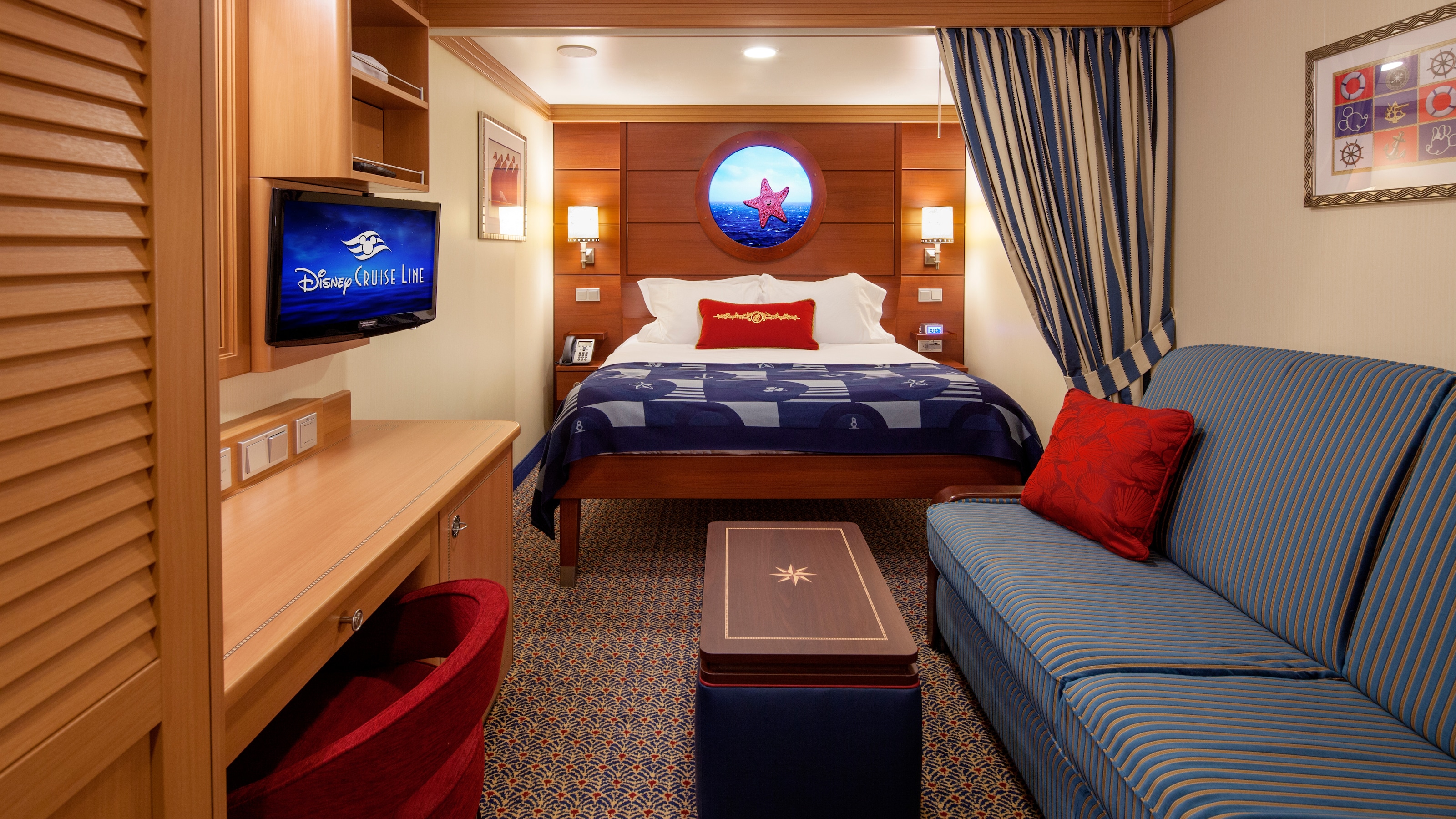 A Disney Cruise Line Standard Inside Stateroom with a desk, a TV, a bed, a virtual porthole, a sleeper sofa and a low table