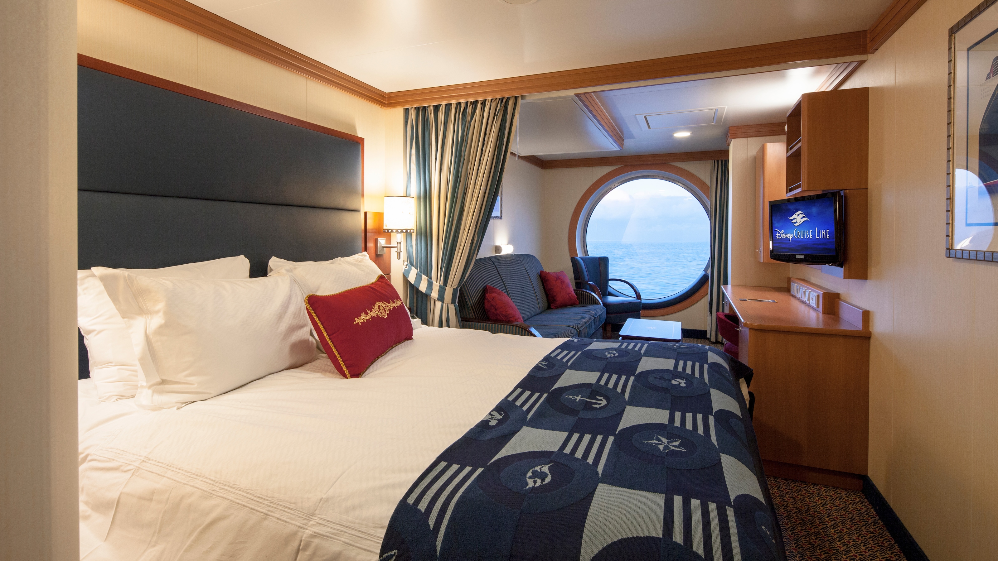 A Disney Cruise Line Deluxe Family Oceanview Stateroom with a queen bed, a sofa, a porthole with an ocean view, storage shelves, a desk and a TV