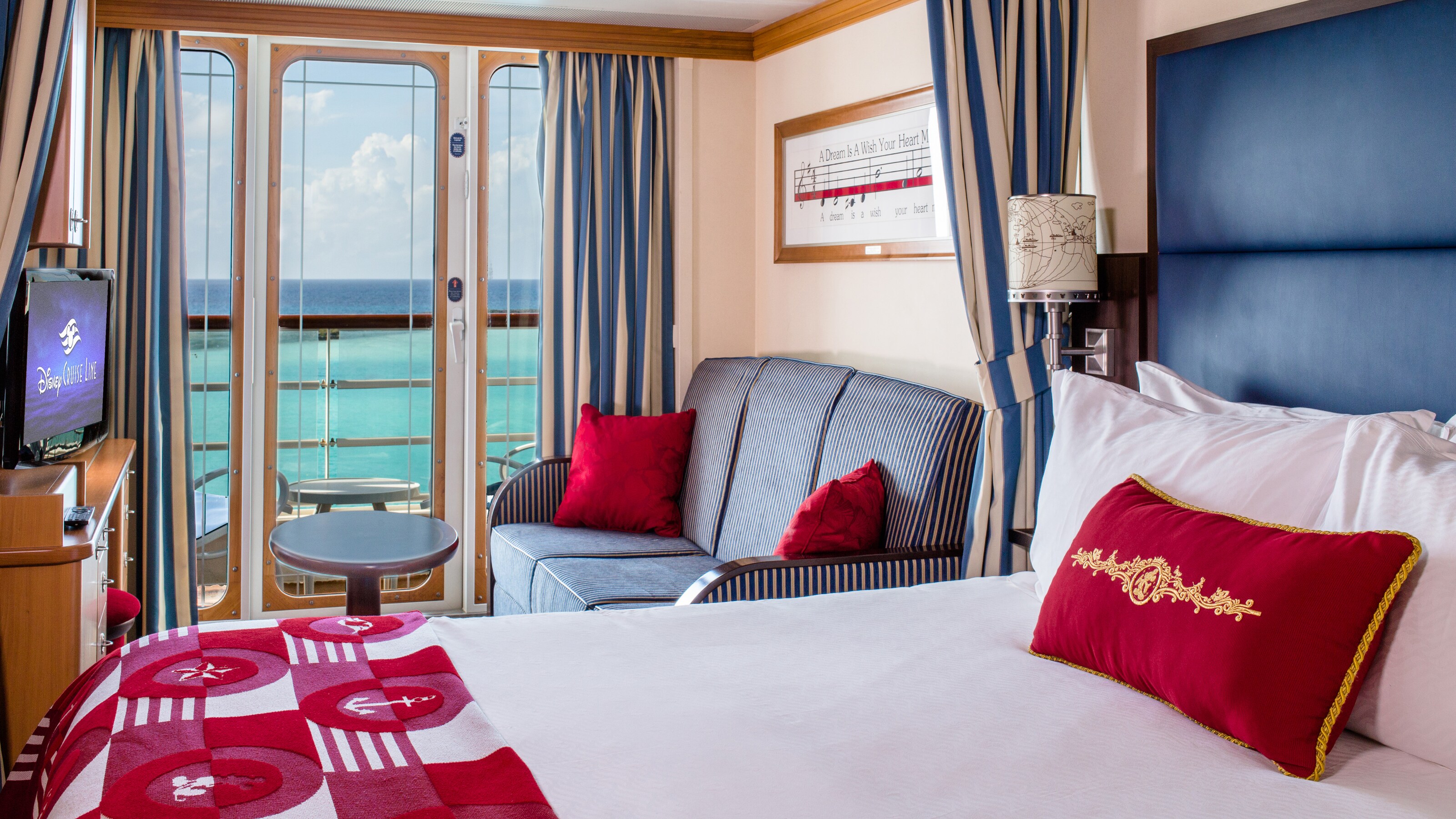 A Disney Cruise Line Oceanview with Verandah Stateroom with a bed, sofa, table, desk, TV and a verandah facing the ocean