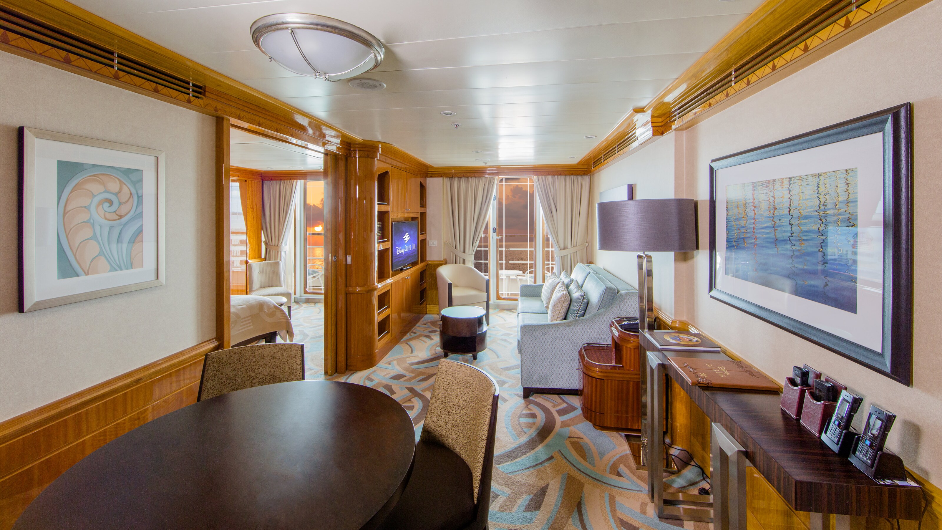 A stateroom suite with a table, couch, lamp, TV and verandah