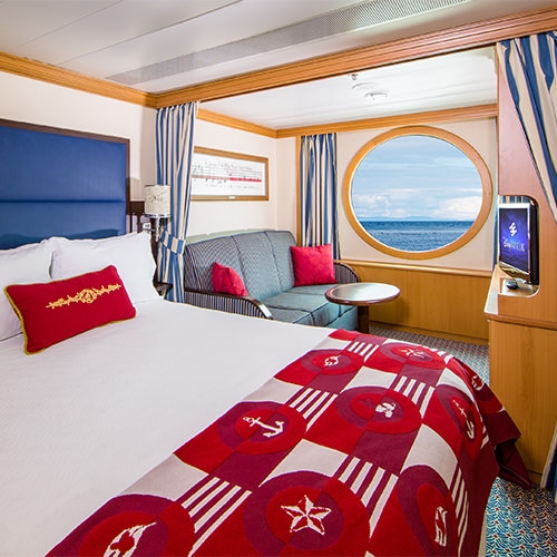 A stateroom with a queen bed, sofa, TV and porthole window 