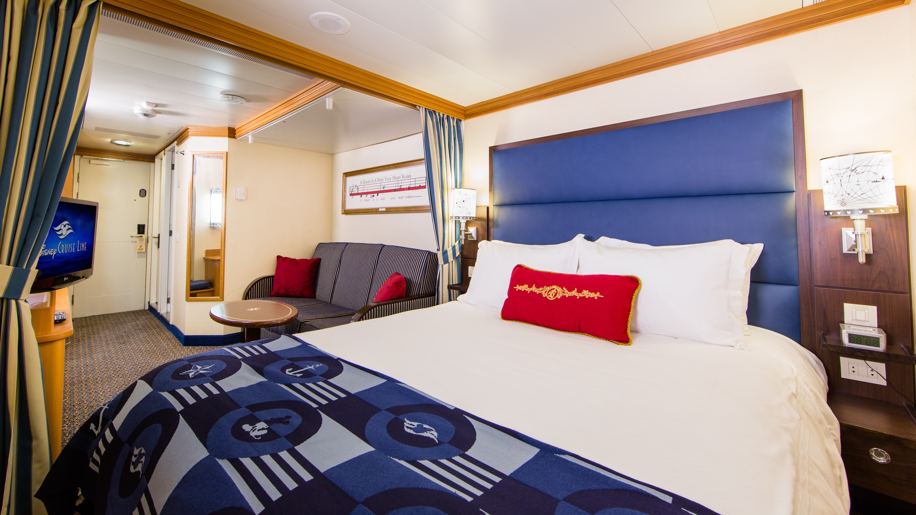 A stateroom with a queen bed, 2 lamps, a privacy curtain, a TV, a sofa and a full length mirror 