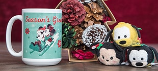 A Season's Greetings coffee mug featuring Mickey and Minnie sledding, a box of pine cones, and three plush toys including Pluto, Mickey, and Minnie.