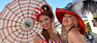 Two women produce happy smiles for a picture, one holds a striped parasol and wears a matching bow on her head, and the other sports a fun red hat.