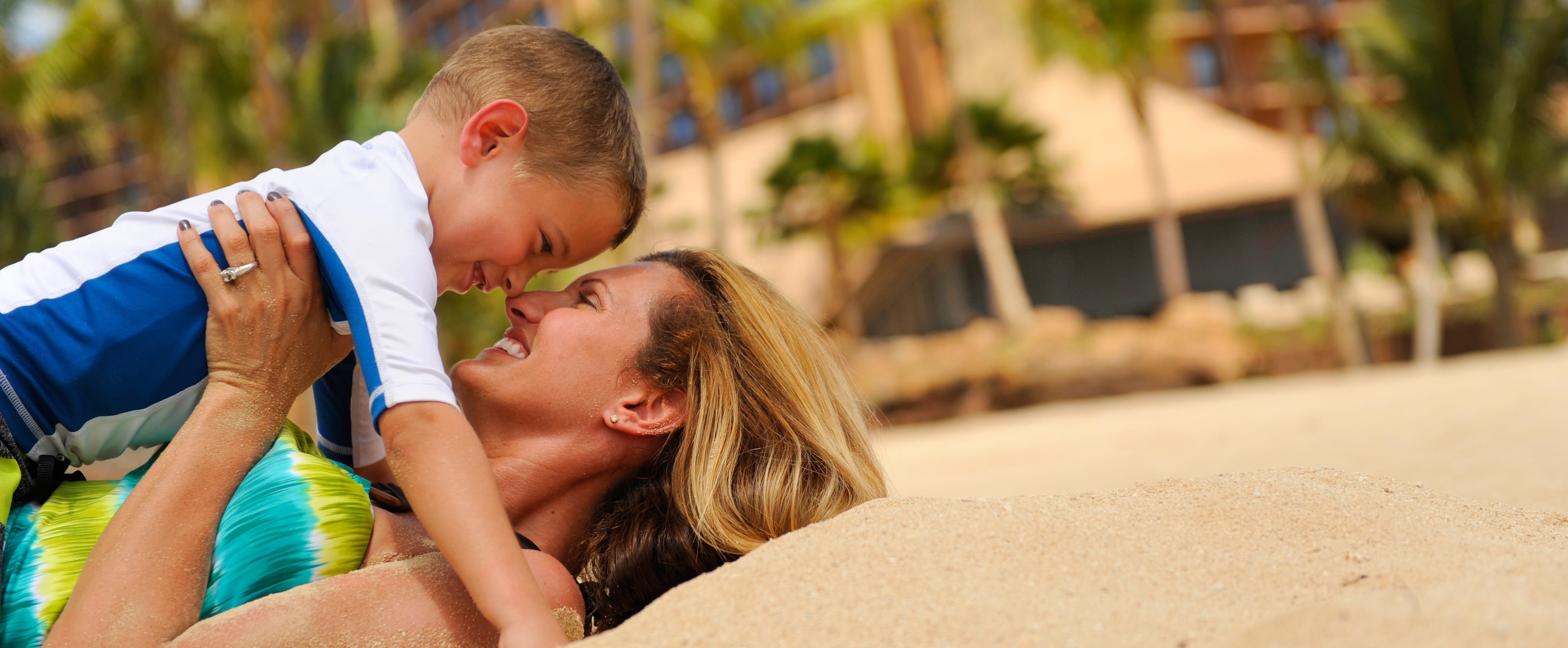 ビーチに仰向けになり、抱っこした小さな息子の鼻に自分の鼻をつけて微笑む母親