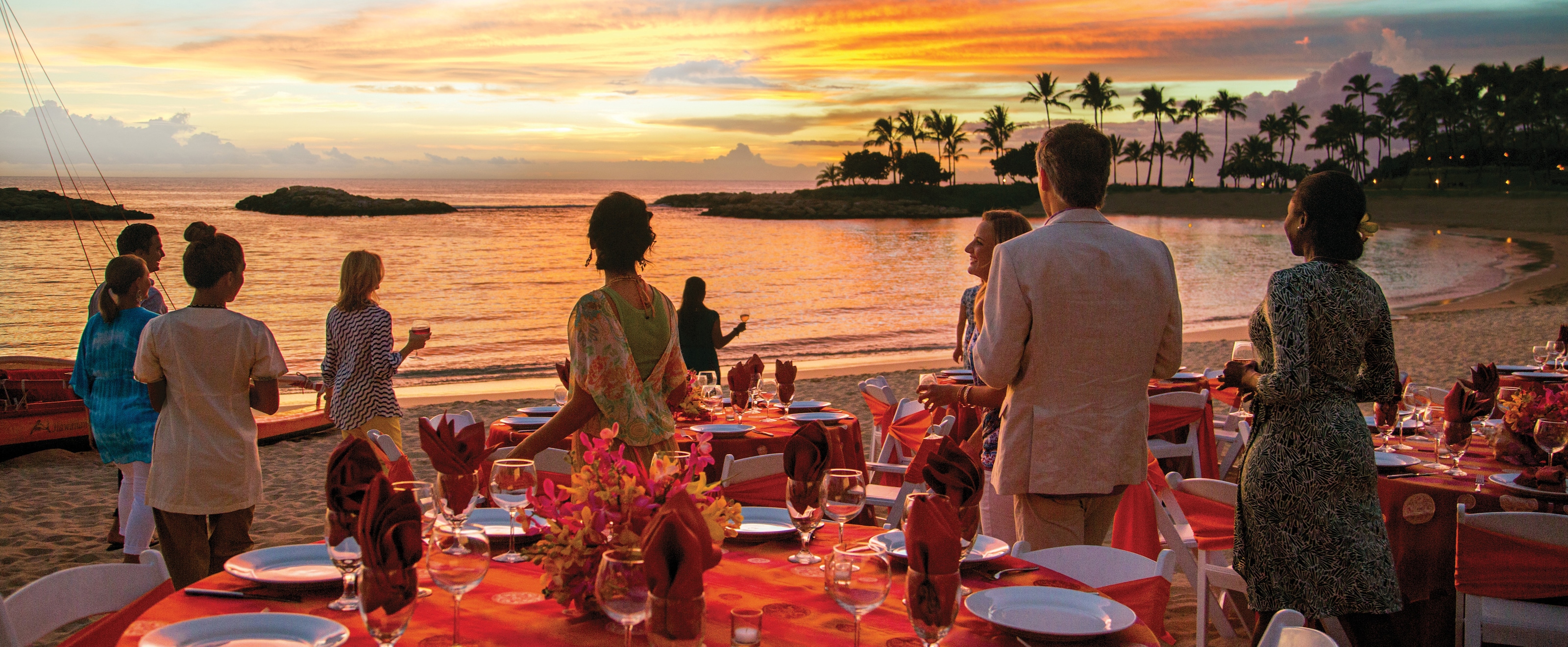 ラグーンにかかる色鮮やかな夕焼けに見とれて立ち止まる海辺のパーティ参加者