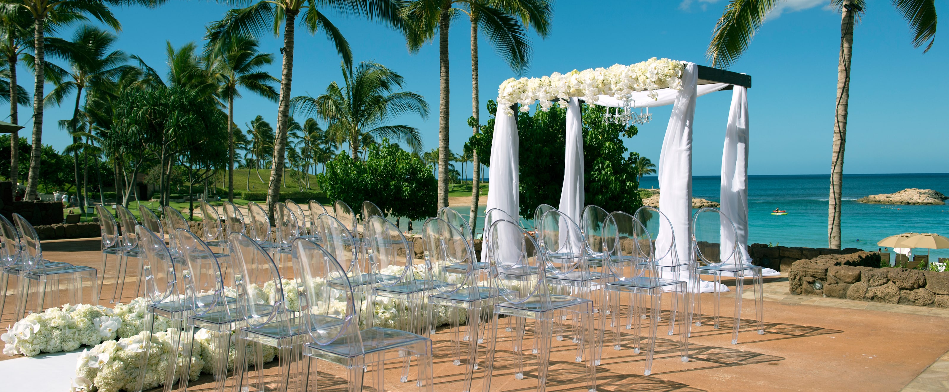 Rows of transparent chairs line an aisle leading to a seaside altar draped in white fabric and flowers