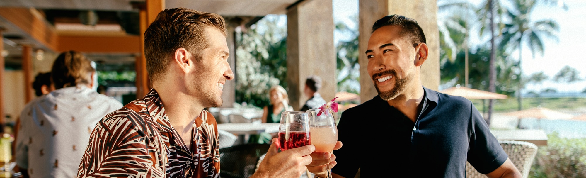 Two smiling young men sitting at a patio table clink their beverage glasses