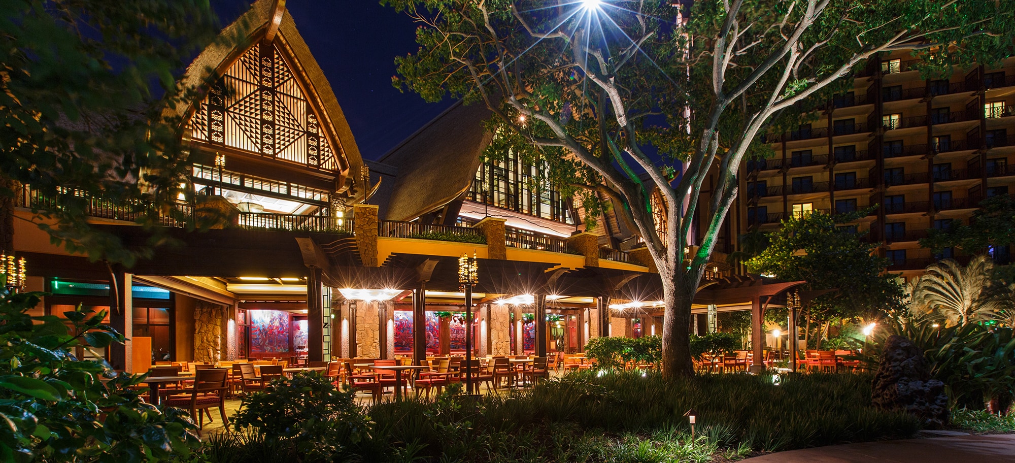 The exterior of Makahiki at night, with a lit-up covered patio and twin peaked roofs with native designs
