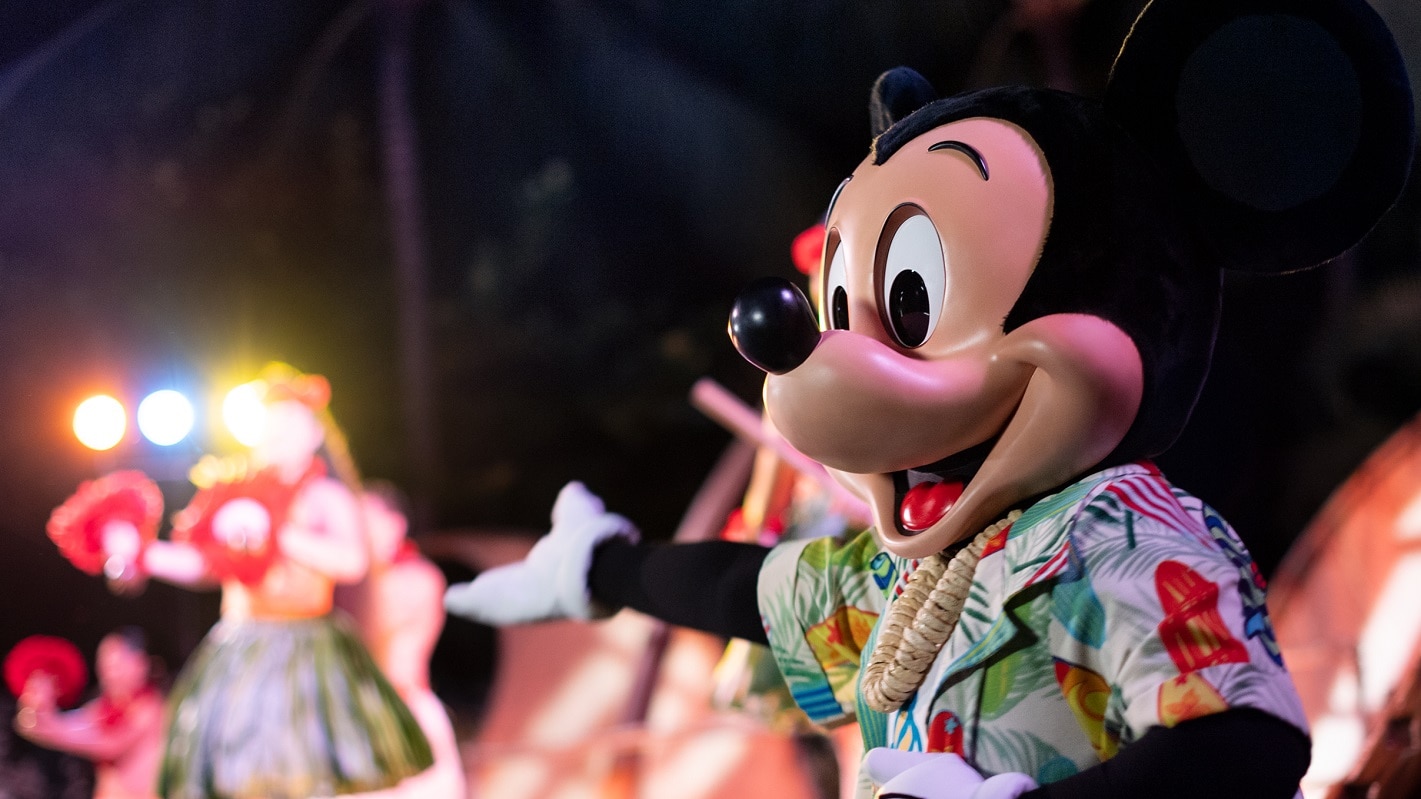 Mickey Mouse, wearing a Hawaiian shirt and a shell lei, joins performers on stage at night