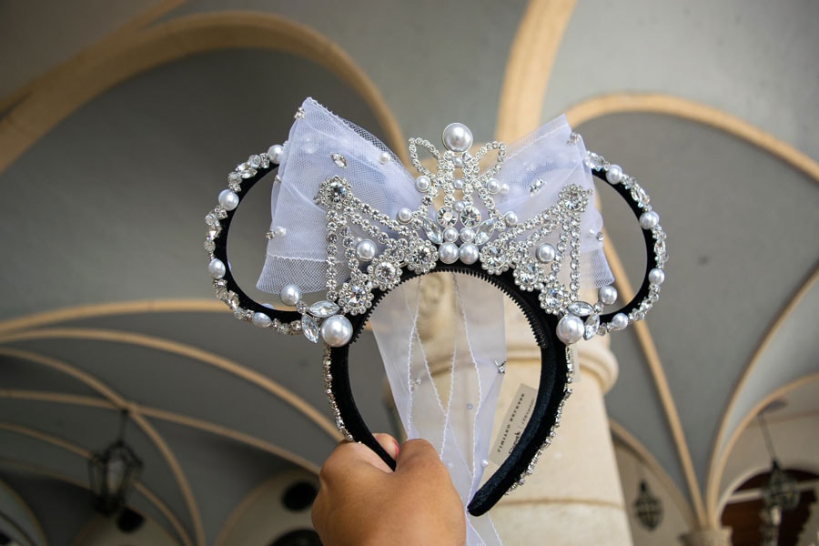 Hand holding the Minnie Mouse  Black Velvet ear headband featuring a combination of pronged faceted glass crystals, acrylic crystals, and synthetic pearls hand-stitched onto the headband and ears. A large off-white organza bow and a Silver plated metal tiara adorned with more glass crystals, pearls and beads.