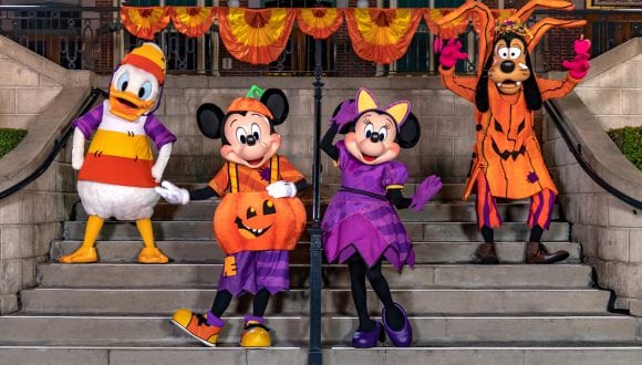 Mickey Mouse as a jaunty jack-o-lantern, Minnie Mouse as a stylish witch, Goofy as a haunted apple tree, Donald Duck as a quirky candy corn