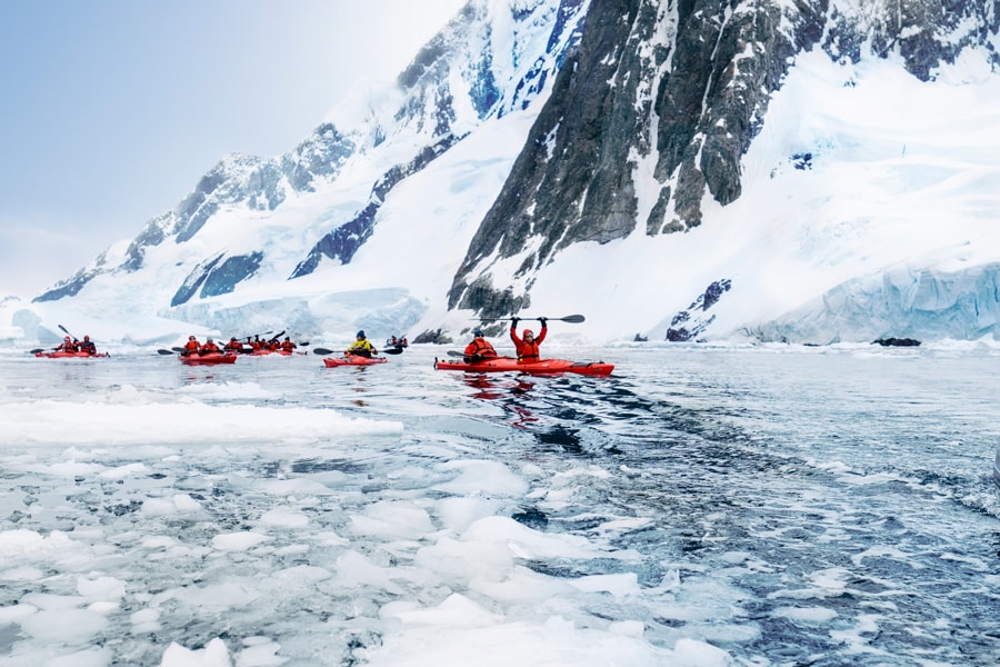 People kayaking in icy water with snow-covered mountains in the background.