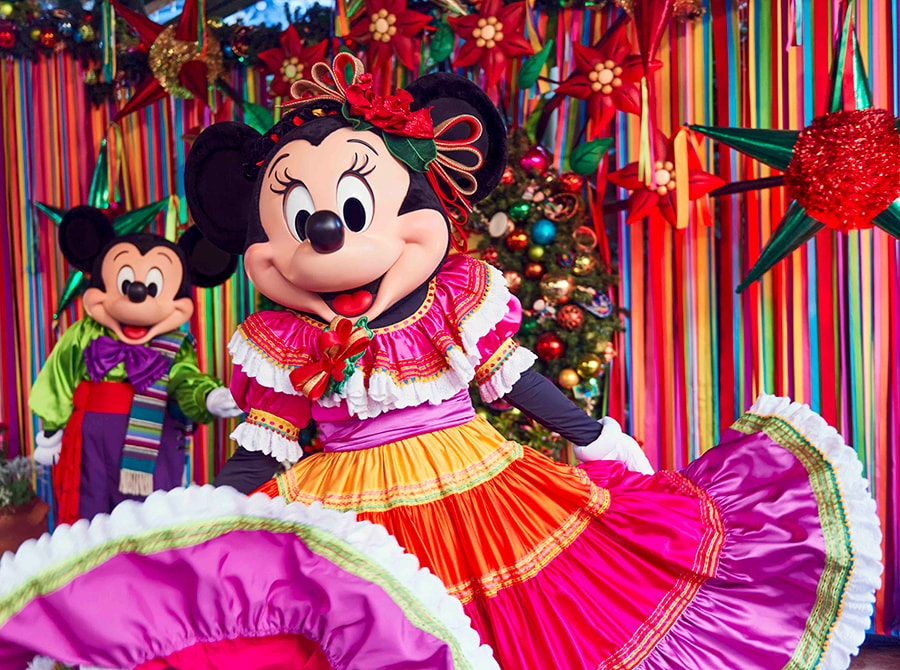 Minnie Mouse fans her skirt in front of Viva Navidad background, with Mickey Mouse slightly blurred in the background.