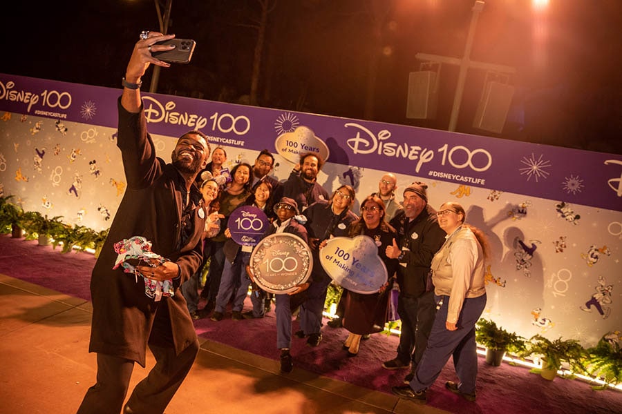 Cast members pose for a selfie in front of a Disney100 photo backdrop