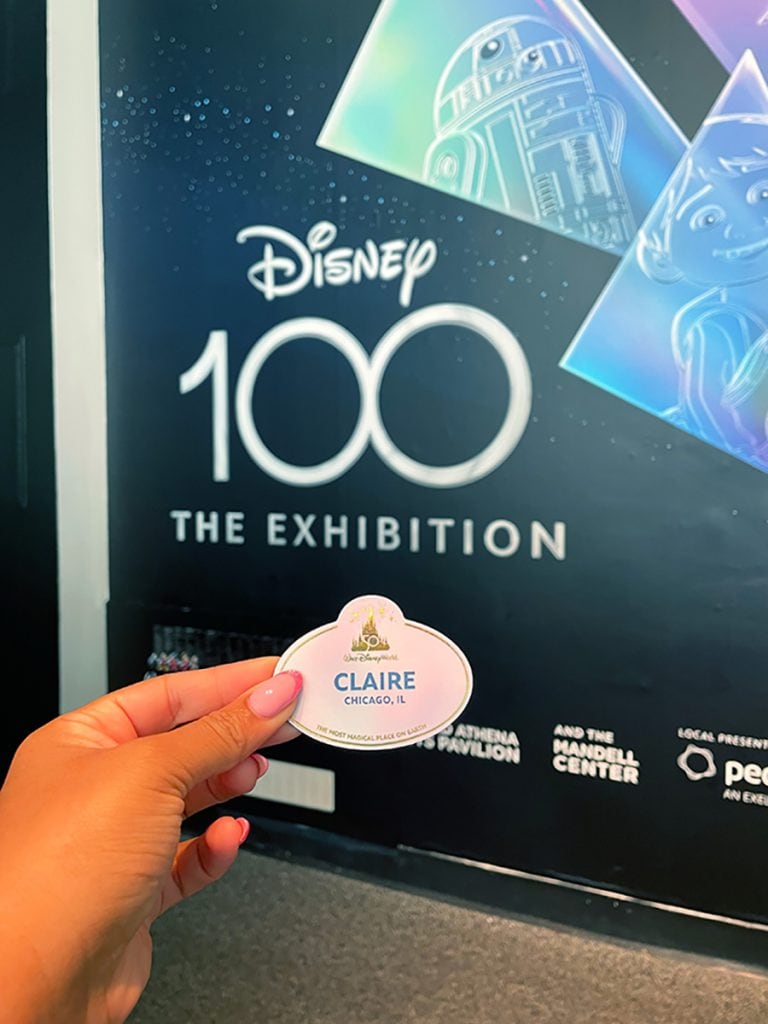 Claire's Walt Disney World nametag held in front of the Disney100 exhibition poster