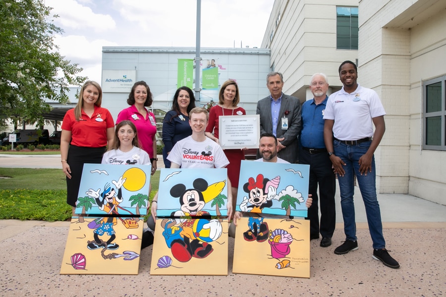 Group of cast members holding up their completed paintings featuring Mickey Mouse, Minnie Mouse and Goofy