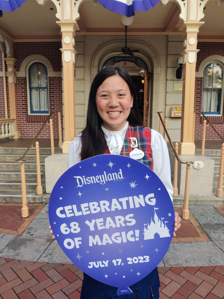 A cast member, Cherilyn, smiles holding a balloon-shaped prop reading "Celebrating 68 Years of Magic - July 17, 2023"