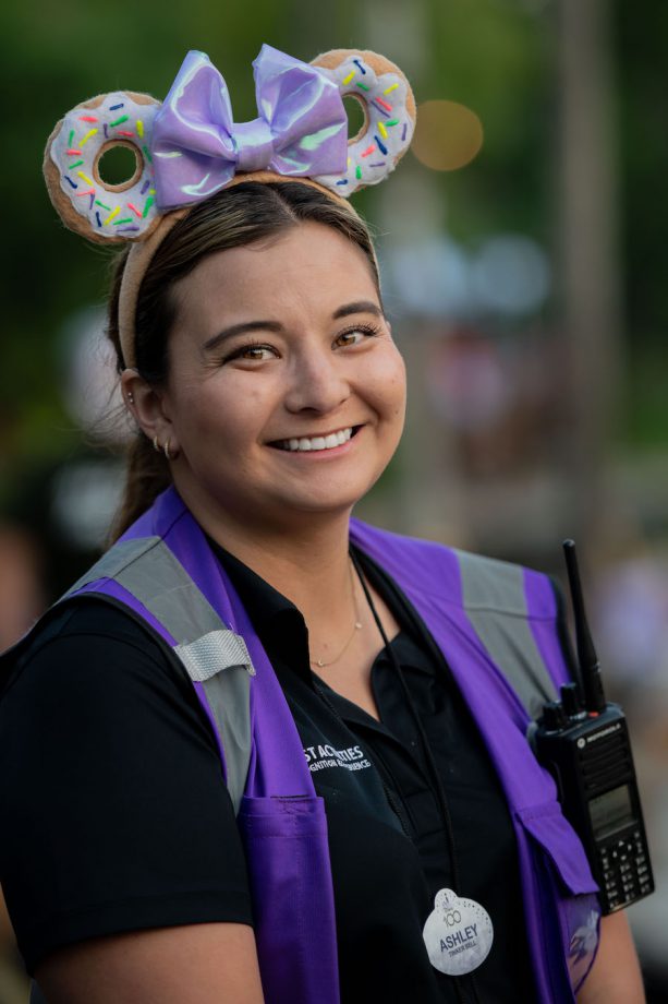 A cast member, Ashley, smiles wearing a purple reflective vest, a pair of donut-themed Minnie ears and a walkie talkie