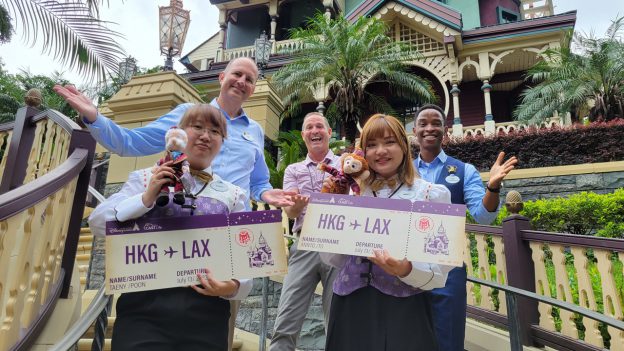 Two cast members holding plane tickets in front of Mystic Manor, with leaders and a Hong Kong Disneyland Ambassador behind them
