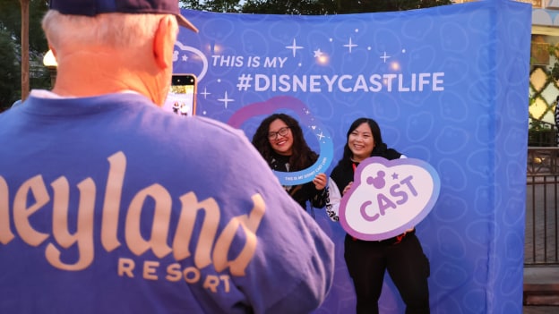 Two cast members holding oversized nametag props pose for a photo in front of a blue backdrop reading "This is My #DisneyCastLife"