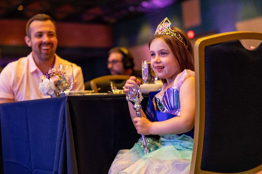 Young female wish kid dressed like Ariel holds a septer while is seated at table with her father looking at her.
