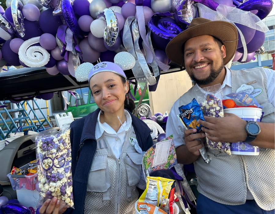 Two PhotoPass cast members stand in front of purple and silver balloons, their arms filled with popcorn, chips and snacks