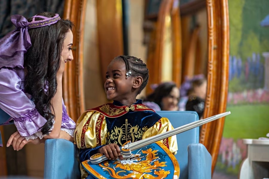 A fairy godmother's apprentice at Bibbidi Bobbidi Boutique talks with a young guest transformed into a brave knight with sword and shield