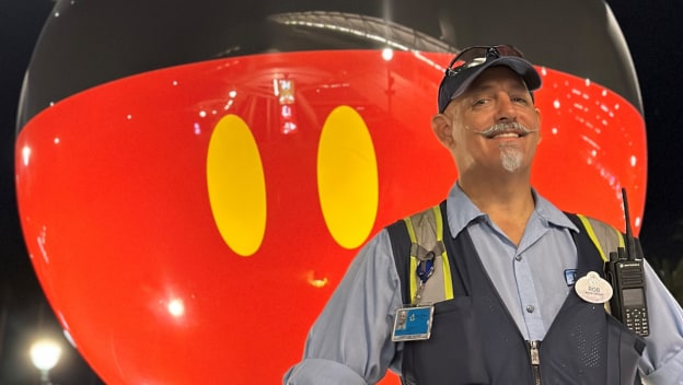 Rob wears his blue custodial outfit and safety vest standing in front of the Mickey Mouse ball in the parking structure plaza at night