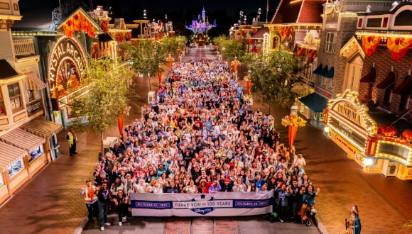 A large group of cast members gathered at nighttime on Main Street, U.S.A. behind a banner reading "Thank You for 100 Years"