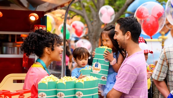 A family gathers around enjoying bags of Disneyland popcorn as guests walk by holding Mickey Mouse balloons