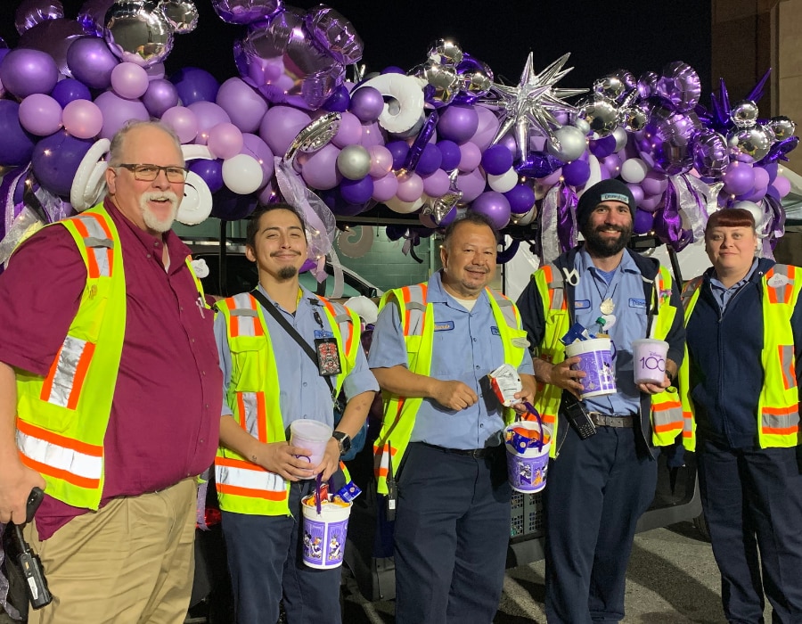 Third-shift cast members wearing reflective safety vests in the early morning hold Disney100 snack buckets and treats in front of a purple balloon backdrop