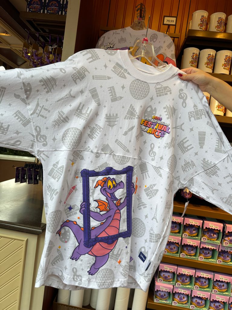 Image of Figment spirit jersey front featuring Figment holding picture frame on a white backdrop featuring icons of art and EPCOT pavilions. 