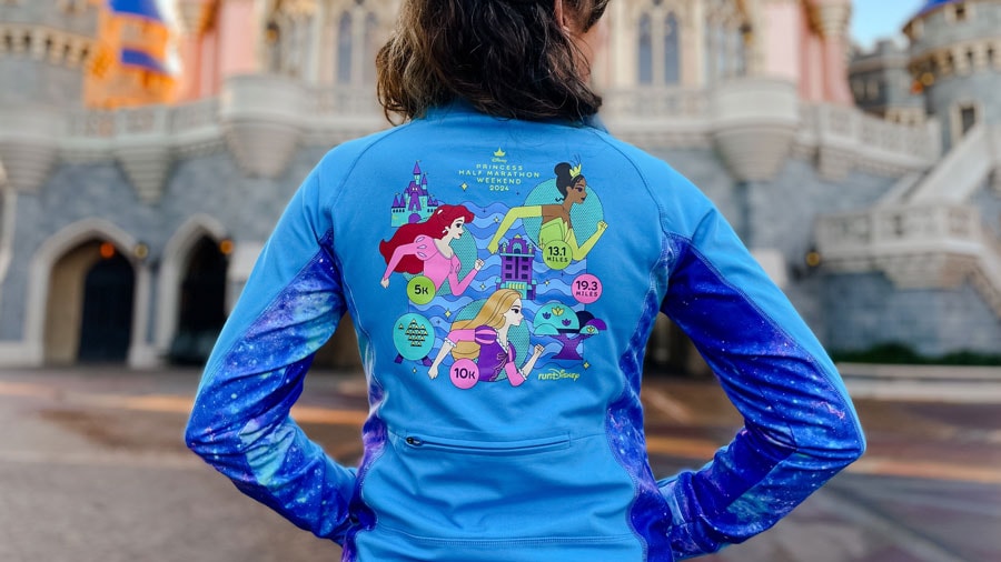 person wearing a jacket themed to princess half marathon weekend 2024 in front of Cinderella's castle