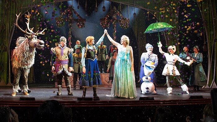 Em uma apresentação ao vivo, Anna e Elsa dão as mãos enquanto outros Personagens de Frozen observam. 