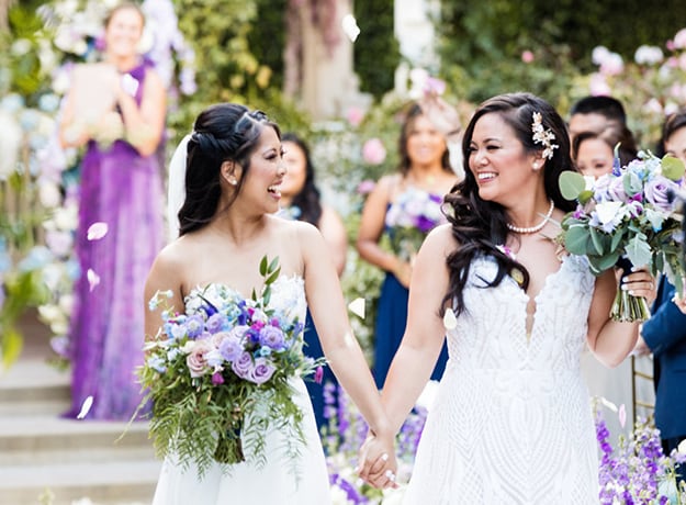 Two women hold hands as they walk down the aisle holding bouquets of flowers as wedding guests look on